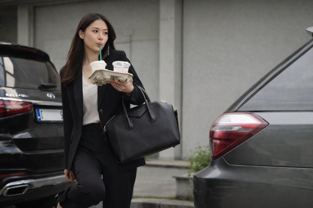 都會女性手持飲品與提袋,穿梭於停車場與車輛之間,象徵通勤接送與多工節奏下的忙碌日常
